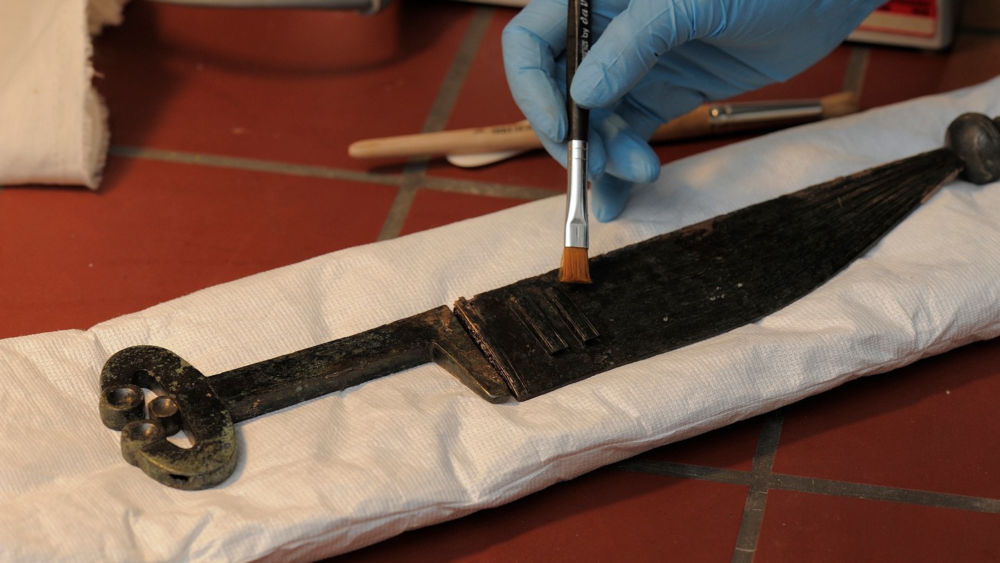 : Gloved hand cleaning an ancient blade with a brush; the blade rests on white padding. (AI-generated alt-text, generated with GPT-4.1-mini)