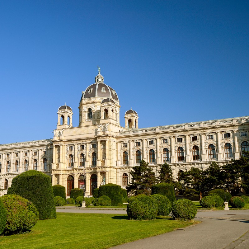 : Historisches Museumsgebäude mit Kuppel und flankierenden Türmen, umgeben von gepflegten Rasenflächen und geschnittenen Büschen unter blauem Himmel. (KI-generierter Alt-Text, erstellt mit GPT-4.1-mini)