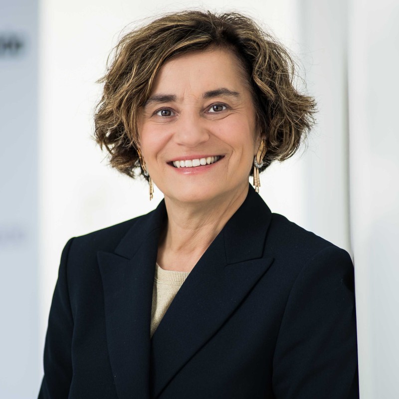 : Woman with curly brown hair in a dark blue blazer, light top, and subtle earrings, smiling at the camera. (AI-generated alt-text, generated with GPT-4.1-mini)