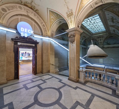 : Entrance area to the exhibition room 'Deck 50' with marble floor, open double doors and modern light installation on walls and ceiling. (AI-generated alt-text, generated with GPT-4.1-mini)