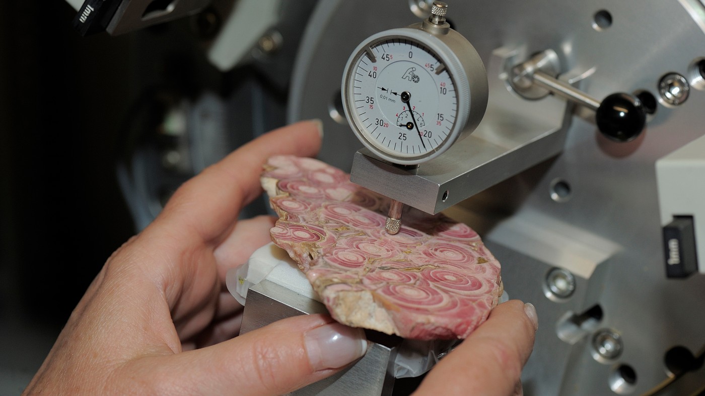 : Close-up of hands holding a polished rock slab with round pink-grey patterns under a measuring device. (AI-generated alt-text, generated with GPT-4.1-mini)