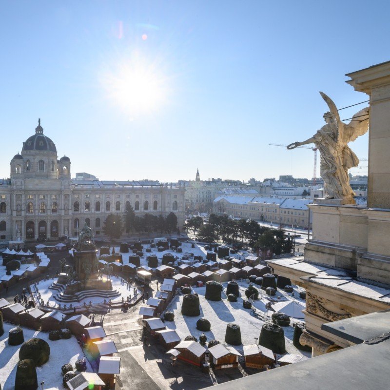 : Blick von einem verschneiten Dach auf das Naturhistorische Museum Wien, links Frontansicht des Gebäudes, rechts eine vergoldete Skulptur, im Hof schneebedeckte Buden. (KI-generierter Alt-Text, erstellt mit GPT-4.1-mini)